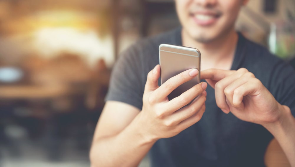 Homem negro com moletom bege mexendo no celular e sorrindo